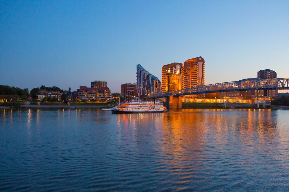 Evening city skyline across a calm river, with a lit bridge and buildings reflecting orange lights on the water.