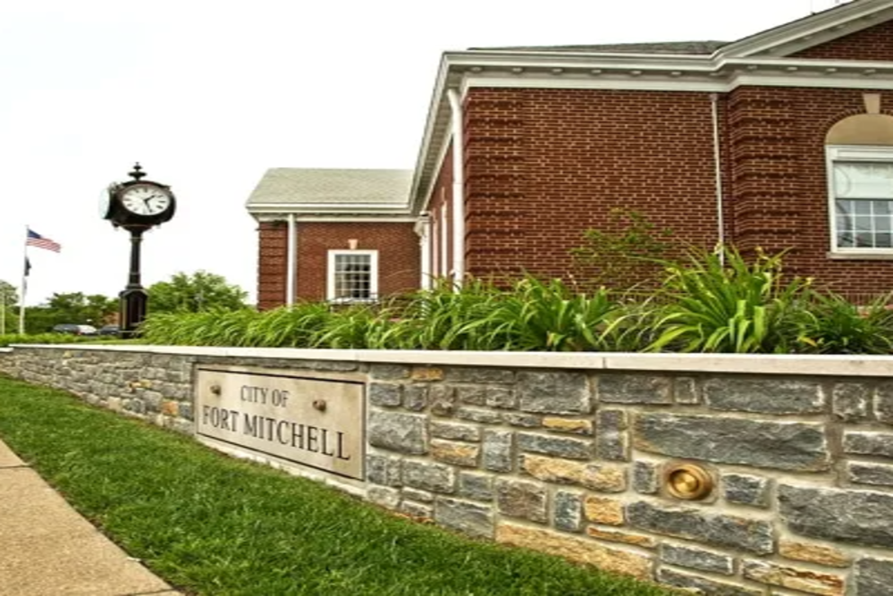 Stone sign on a low wall reading 'City of Fort Mitchell' beside a brick municipal building and a clock tower.