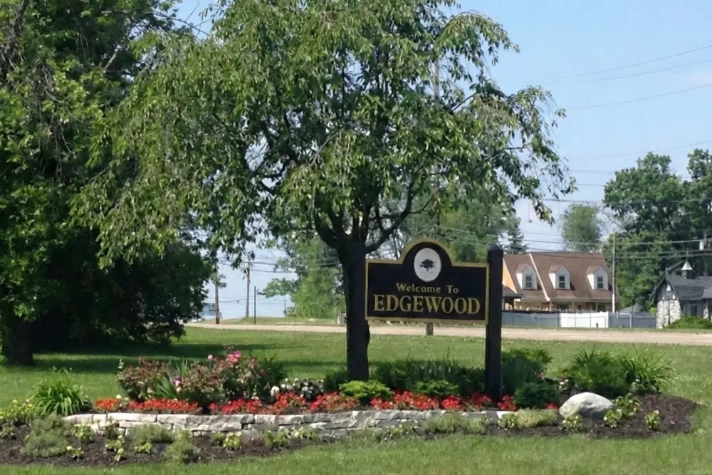Welcome sign reading 'Edgewood' set in a landscaped flower bed with trees and houses in the background.