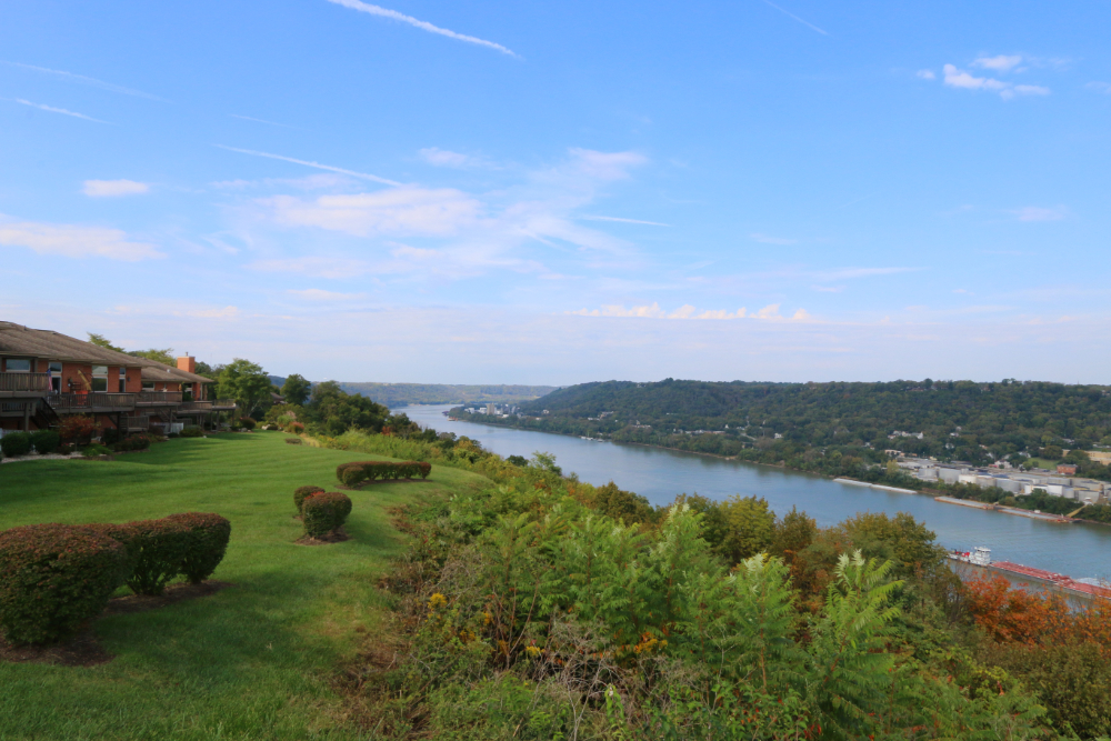 Scenic overlook: green lawn with trimmed hedges on a terraced slope, river valley beyond, and houses on the left under a blue sky.